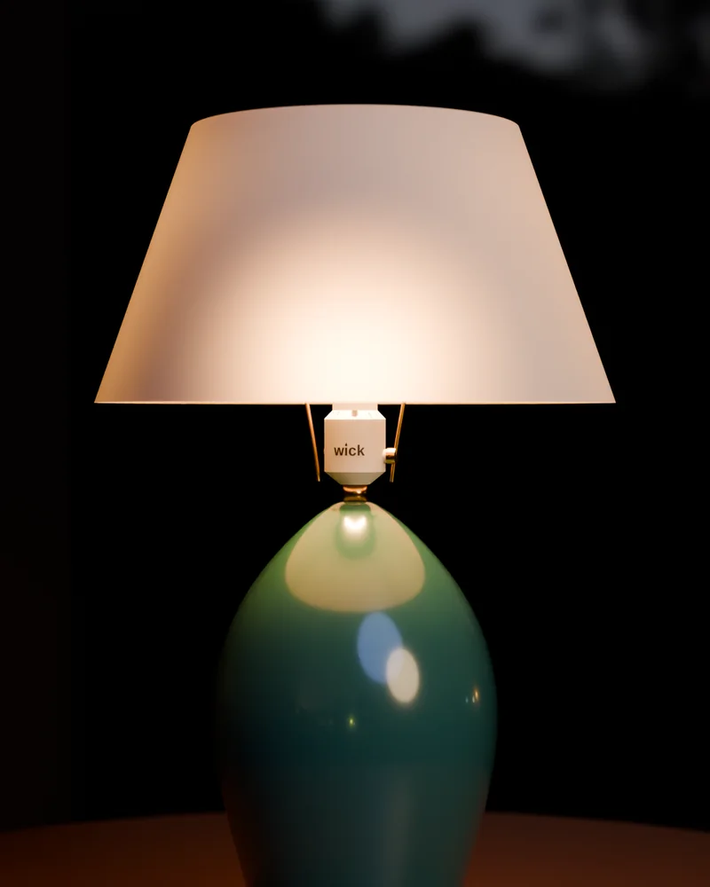 A teal ceramic table lamp with a lit linen shade and a white wick socket beneath it